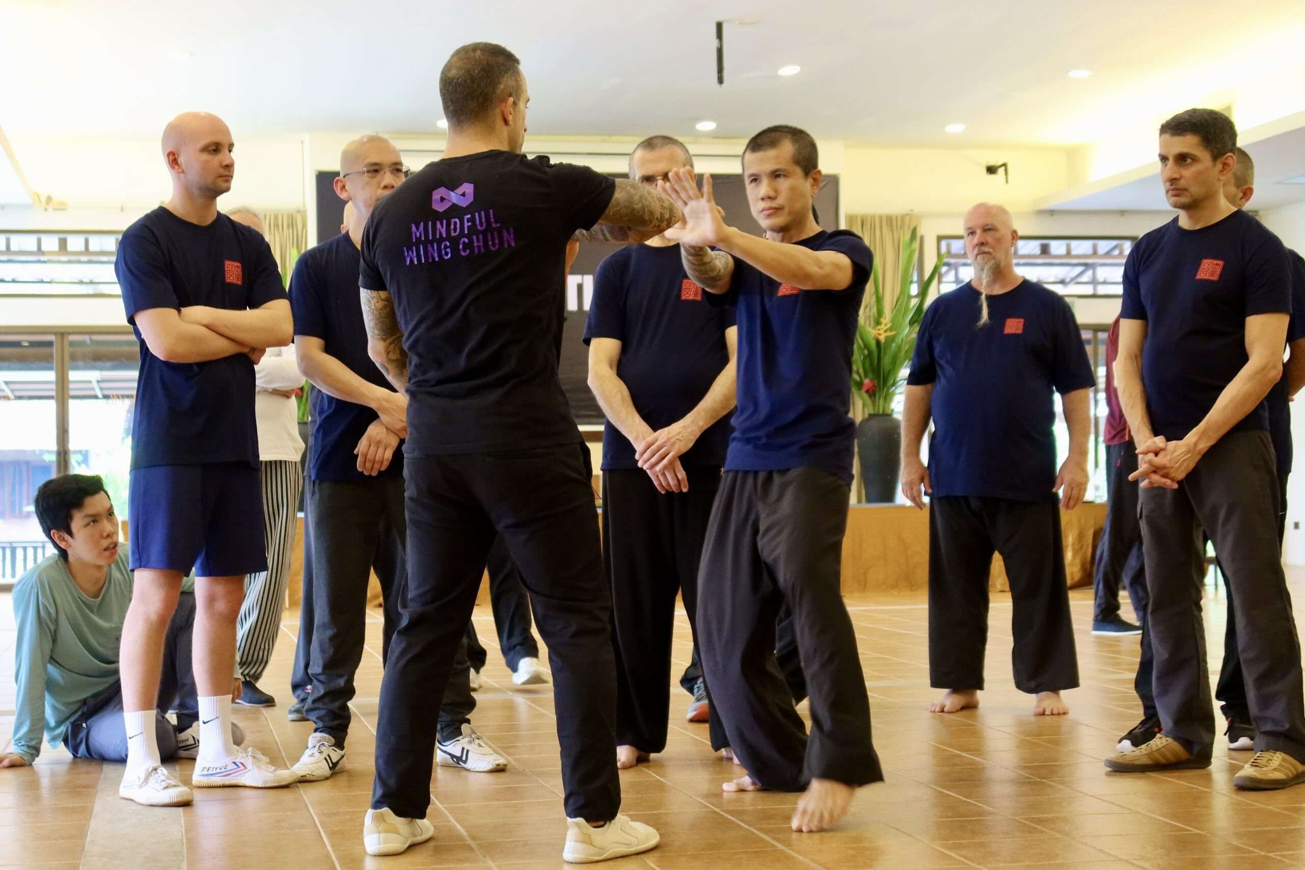 A teacher in black demonstrates on a participant while the group stands in a line watching