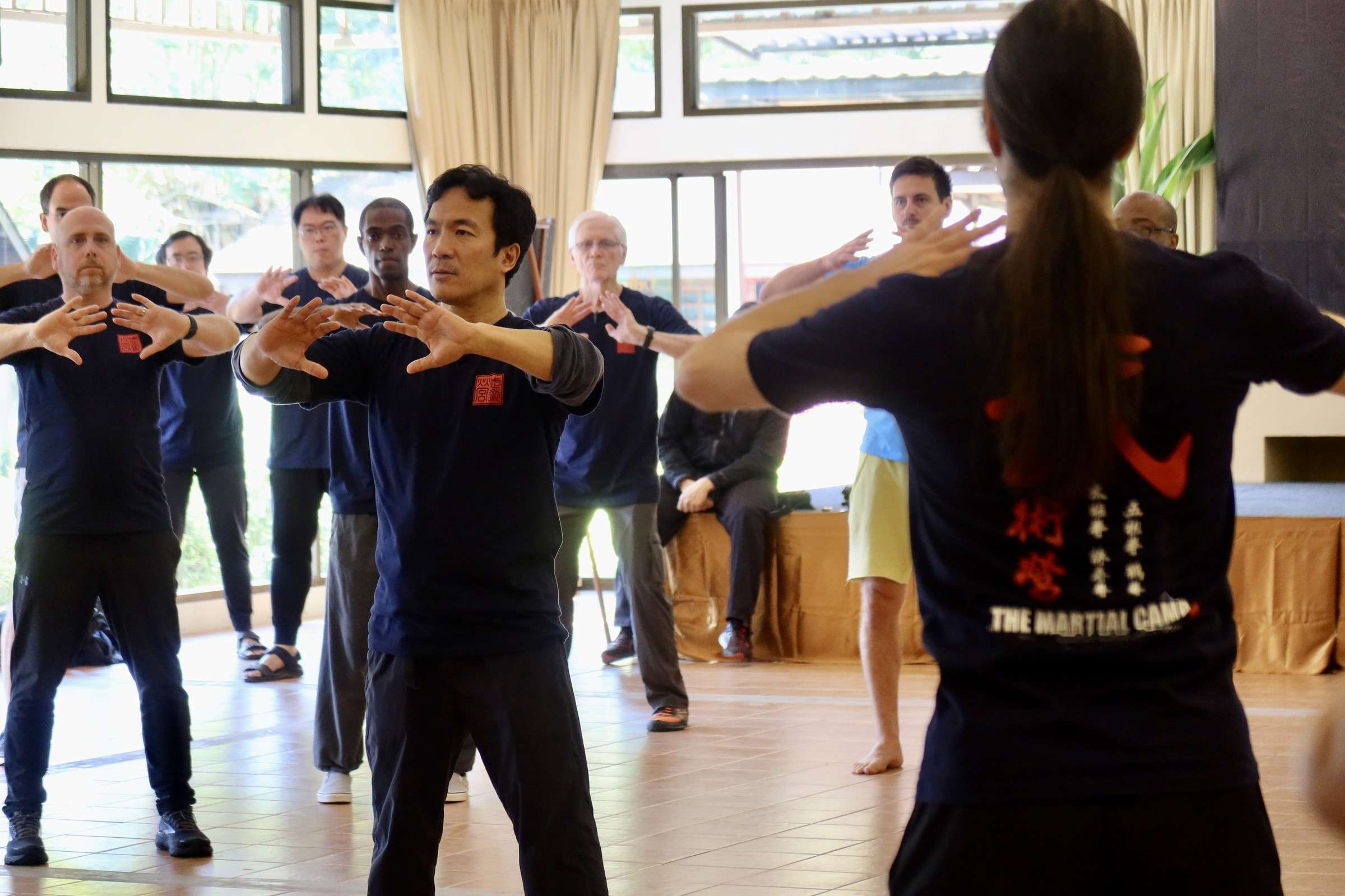 Participants follow a standing exercise together in the training hall, hands raised in front of them