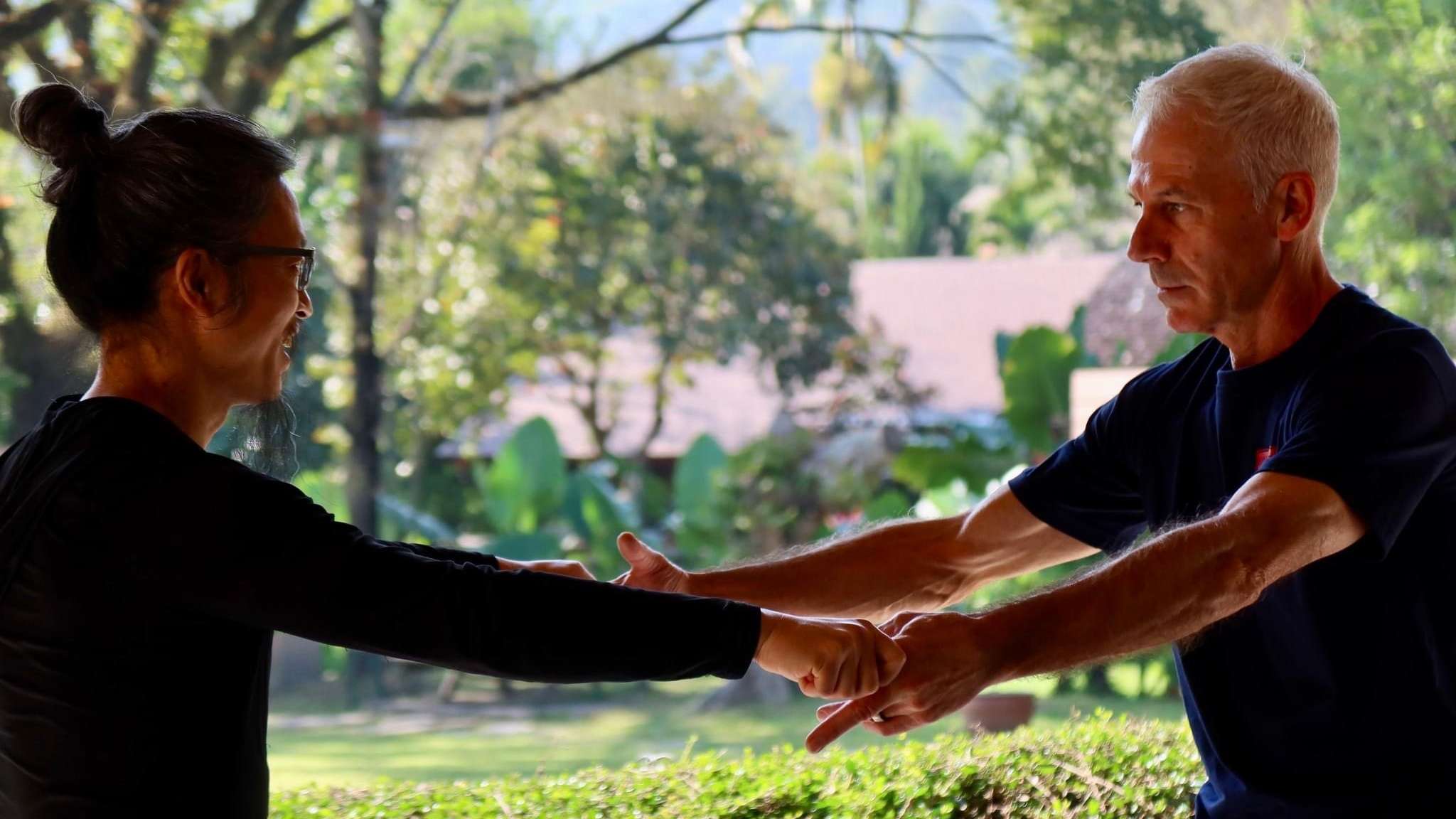 Two practitioners train arm to arm outdoors beside a leafy garden