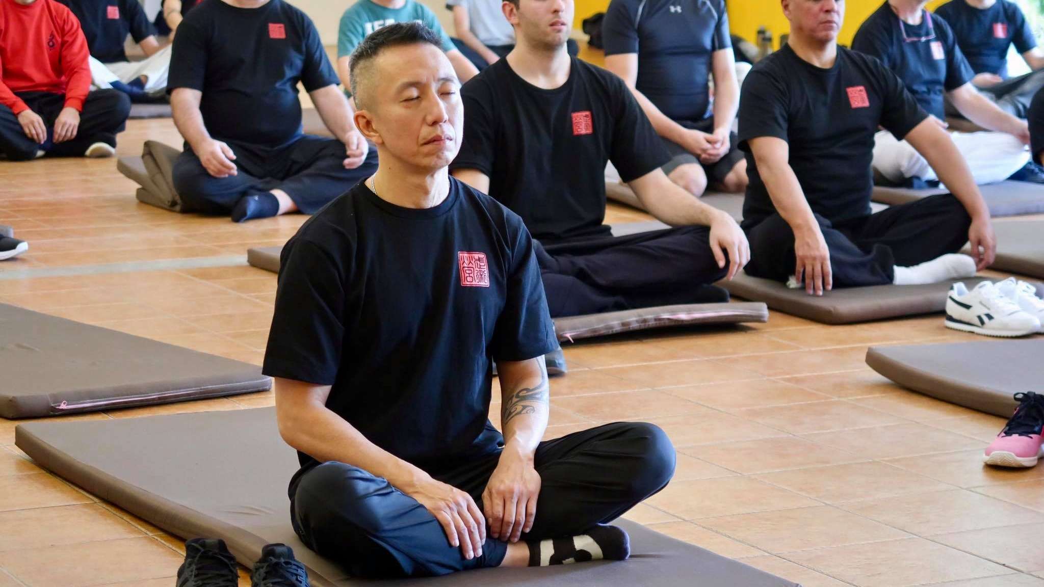 Participants sit cross-legged on mats in the training hall, eyes closed in meditation