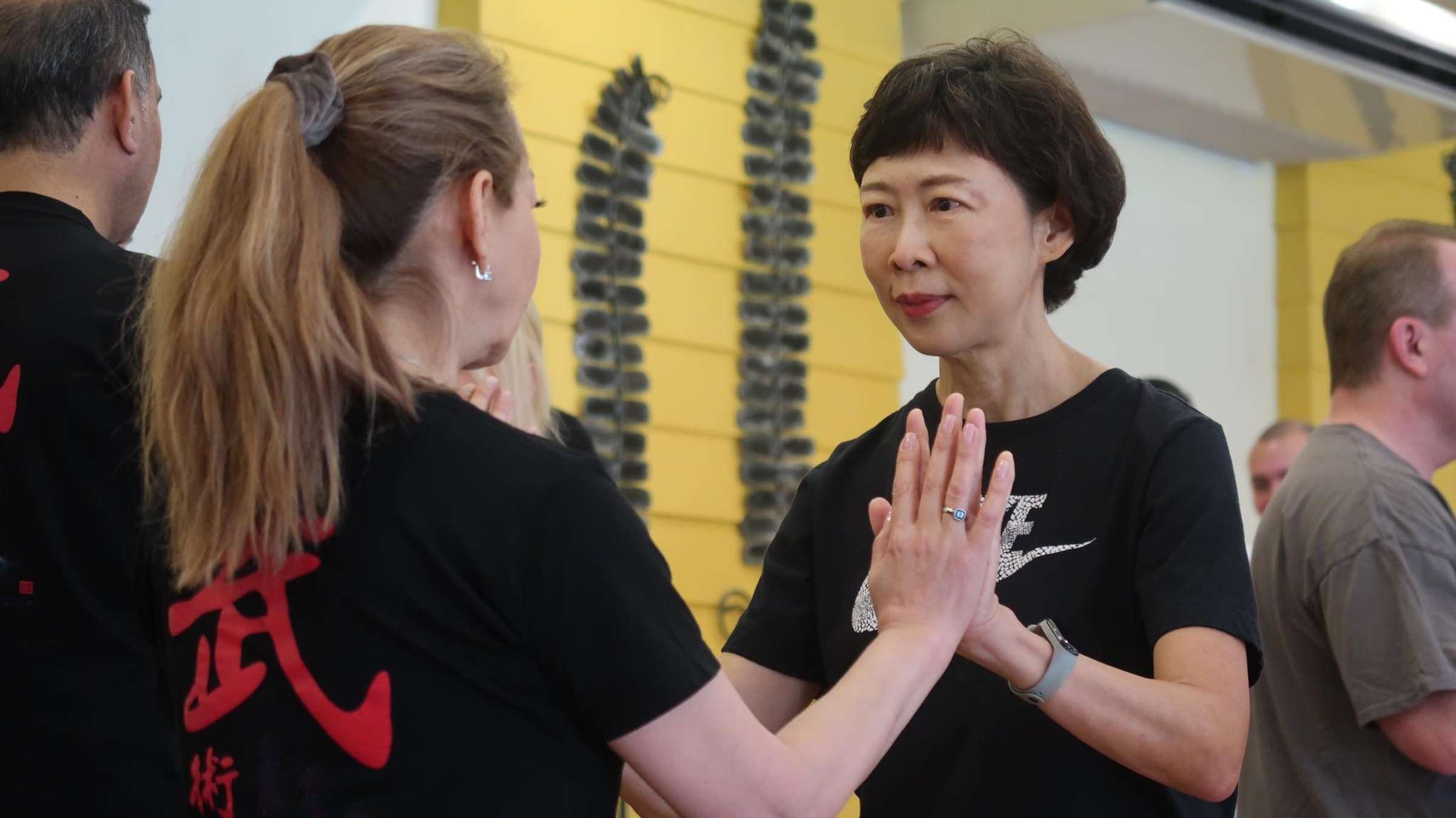 Two women practise a palm-to-palm partner drill in the training hall, one smiling slightly