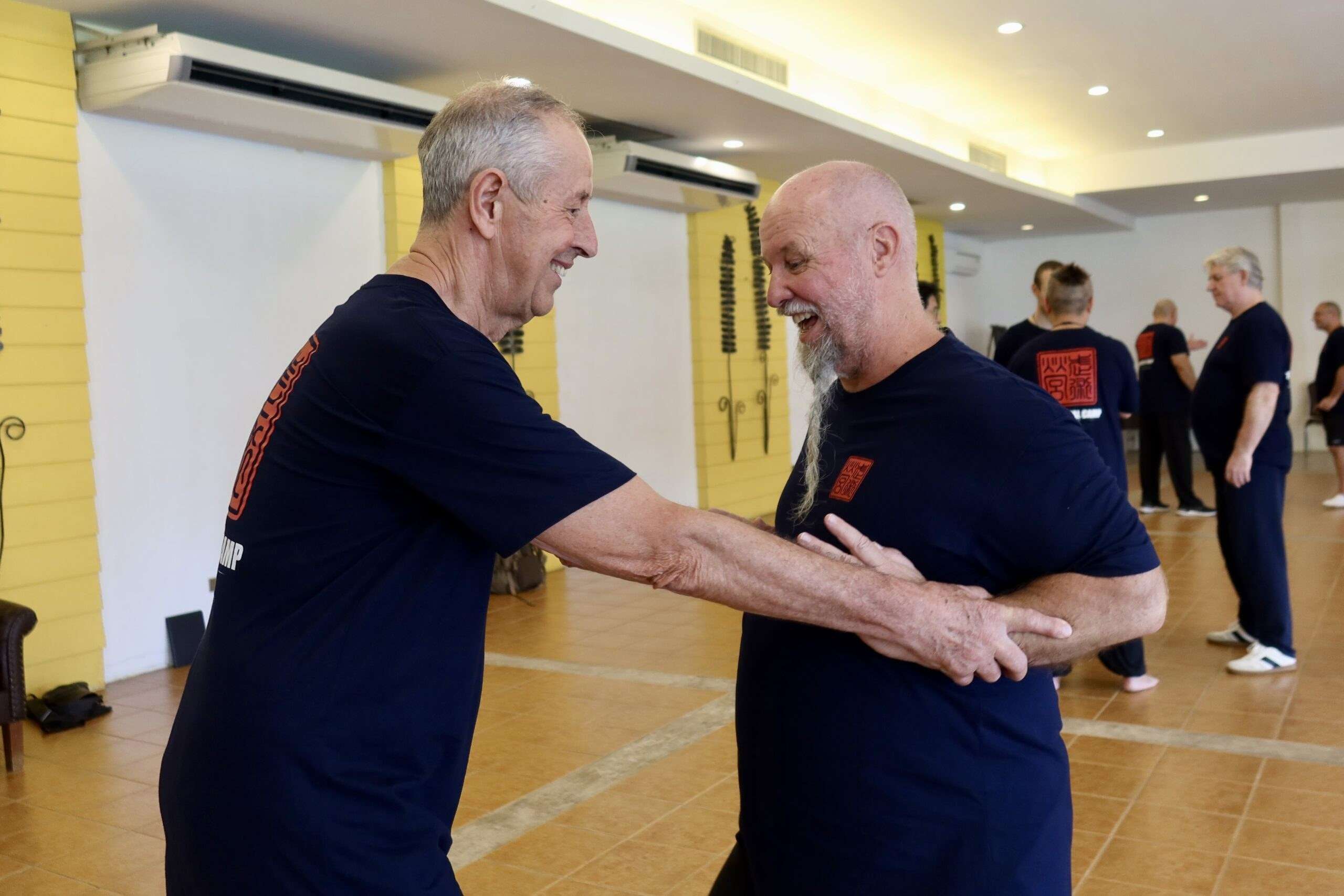 Two older participants laugh together while practising a partner drill in the training hall