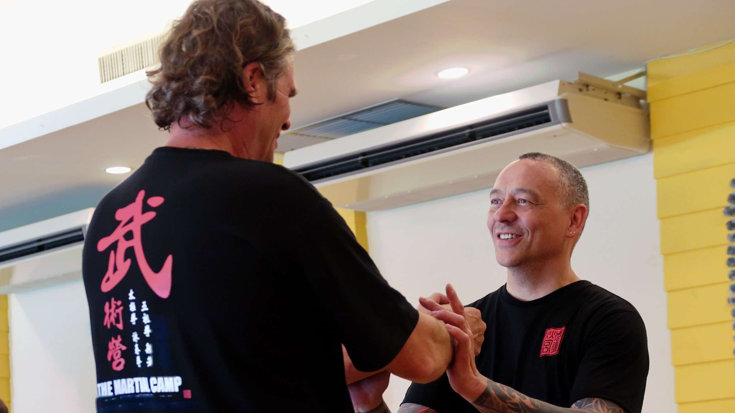 Two men practise a partner drill with hands joined, one smiling broadly at the other