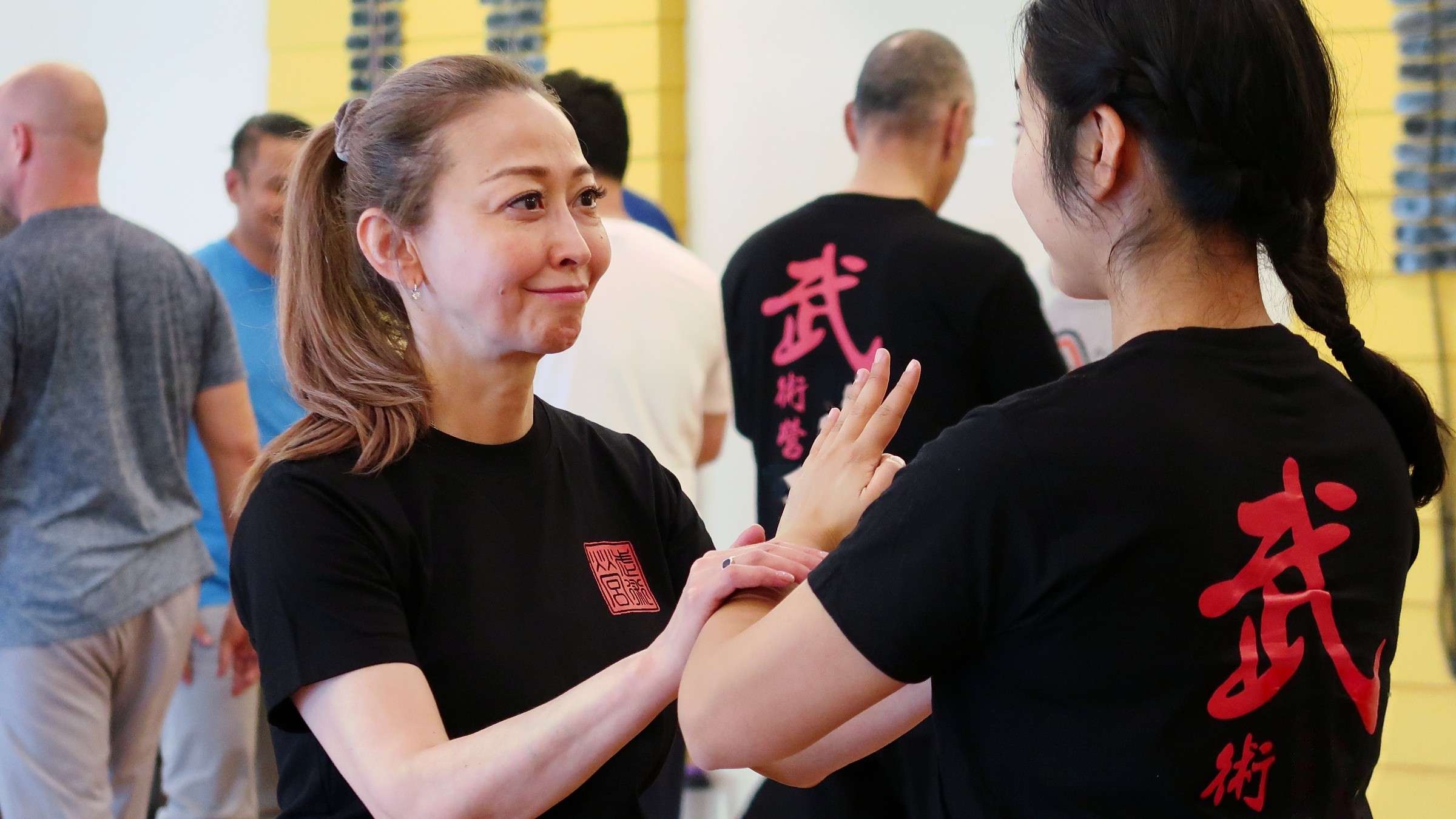 A smiling woman practises a sticky-hands drill with a partner as other participants train behind them