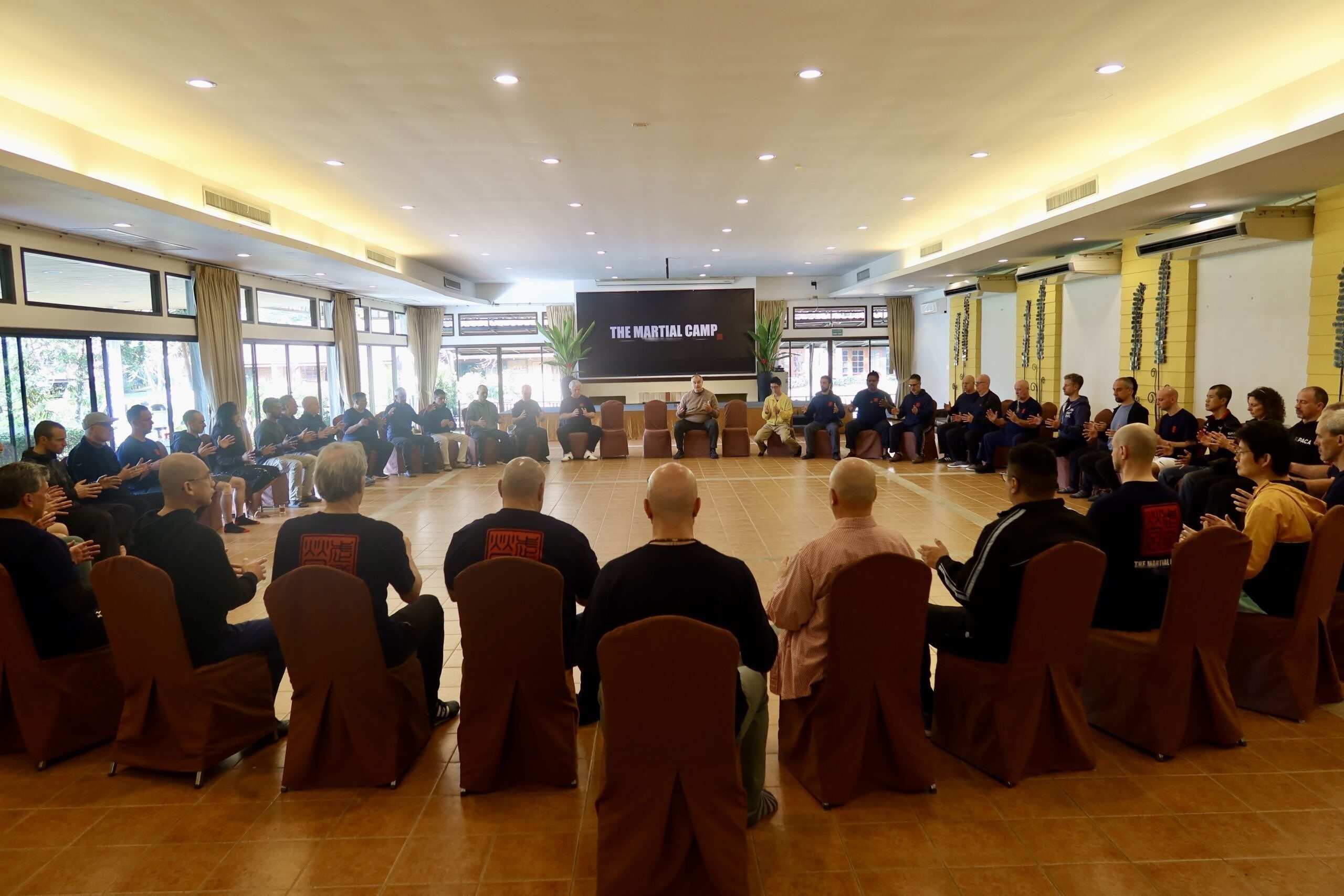 Participants seated in a large circle in the bright training hall, listening to a teacher