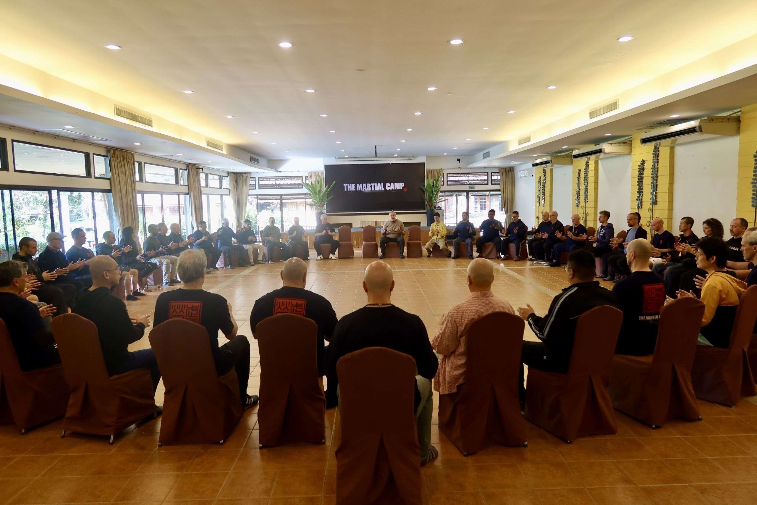 Participants seated in a large circle in the bright training hall, listening to a teacher
